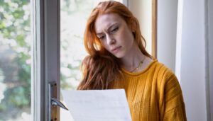 Woman with light skin and bright red hair looking at a document. She has a serious expression.