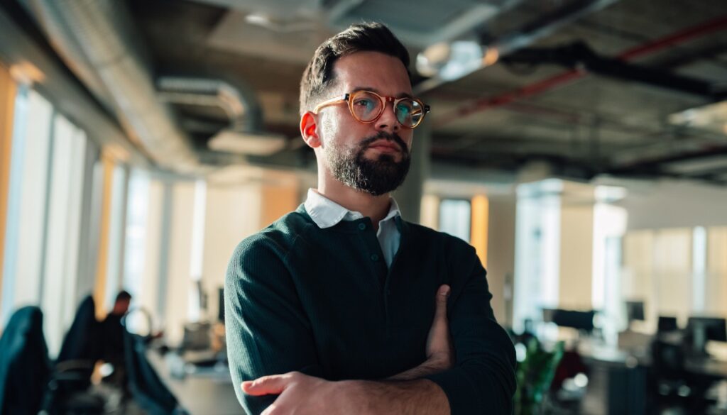 Man in his 30s wearing glasses, looking off screen with his arms crossed. He has a serious expression on his face.